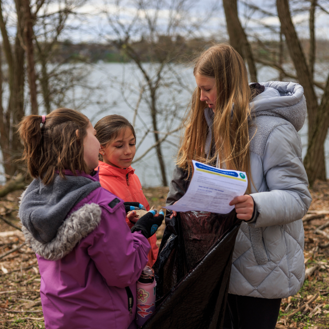 three girl scouts outdoors in front of lake collecting leaves and sticks in trash bag with activity sheet